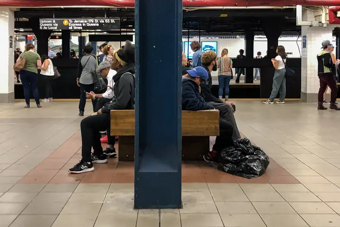 A crowd of people wait on a subway platform, where people sit on a bench including a homeless person.